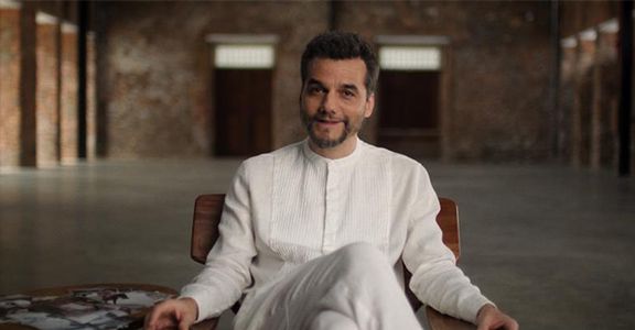Foto de Wagner Moura vestido de branco sentado em uma poltrona.